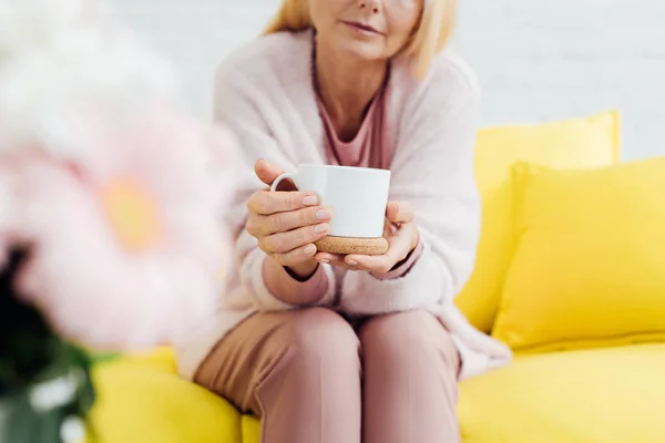 Close up of mature woman sitting with cup of coffee on yellow sofa — Stock Photo