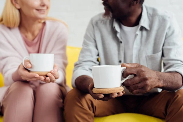 Gros plan d'une femme mûre souriante assise sur un canapé avec un homme afro-américain et buvant du café ensemble — Photo de stock