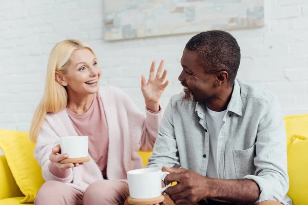 Femme mature émotionnelle assise sur un canapé et parlant avec un homme afro-américain avec des tasses blanches — Photo de stock