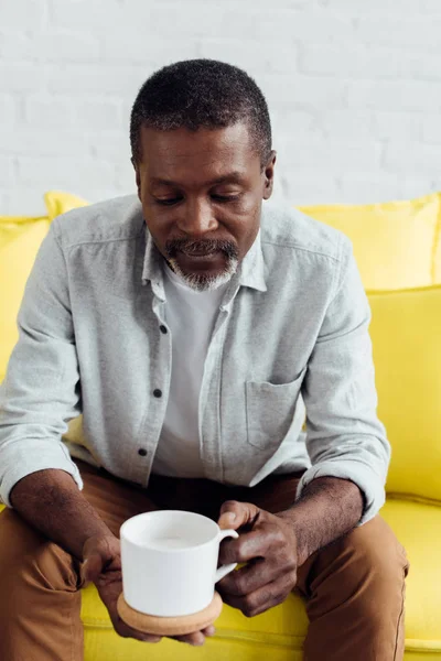 Красивий афроамериканський чоловік сидить на яскраво-жовтому дивані з білою чашкою — стокове фото
