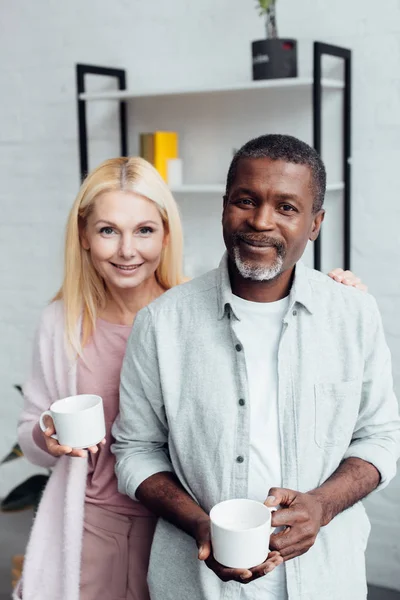 Mujer madura feliz con afroamericano sosteniendo tazas blancas - foto de stock