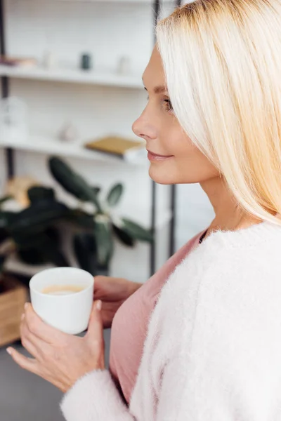 Atractiva sonriente mujer rubia madura sosteniendo taza de café blanco - foto de stock