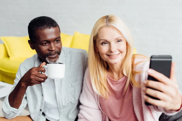 Sonriente madura mujer tomando selfie mientras africano americano hombre bebiendo café - foto de stock