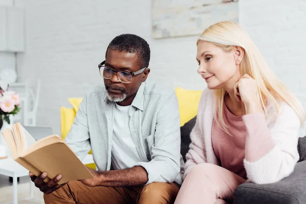 Africano americano hombre en gafas y rubia madura mujer leyendo libro juntos - foto de stock