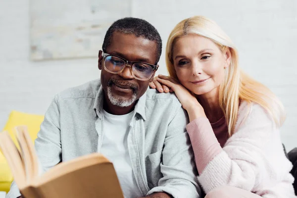 Africano americano hombre en gafas y rubia madura mujer leyendo libro juntos - foto de stock