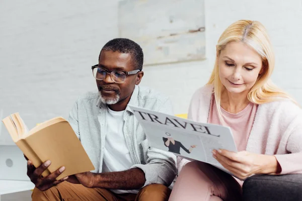 Africano americano uomo in occhiali lettura libro mentre maturo donna lettura viaggio giornale — Foto stock