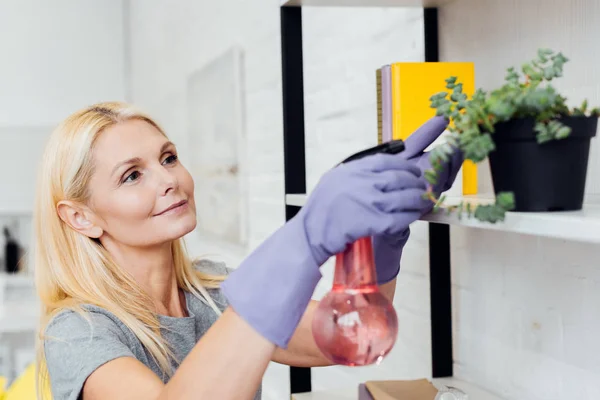 Atractiva mujer madura en guantes de goma pulverización planta verde en rack - foto de stock