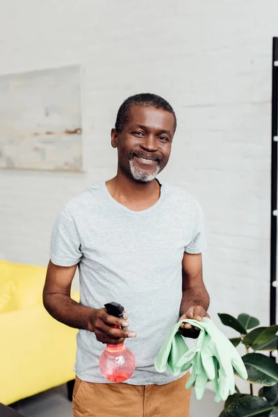 Guapo sonriente afroamericano hombre sosteniendo guantes de goma y spray - foto de stock
