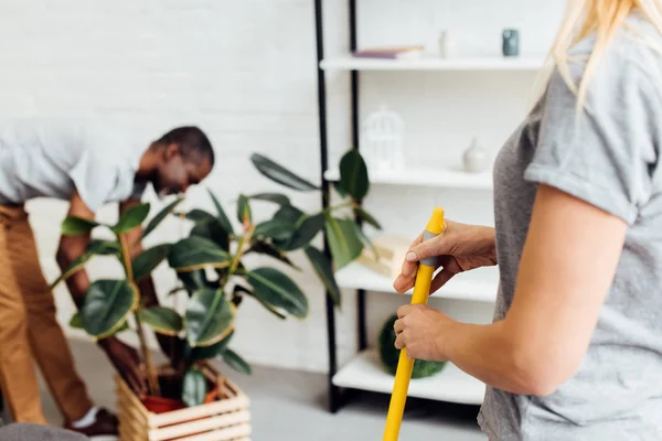 Mujer rubia sosteniendo fregona mientras hombre afroamericano poniendo planta en maceta de madera - foto de stock