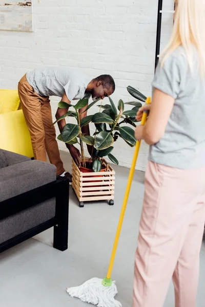 Mujer rubia sosteniendo fregona mientras hombre afroamericano poniendo planta en maceta de madera - foto de stock