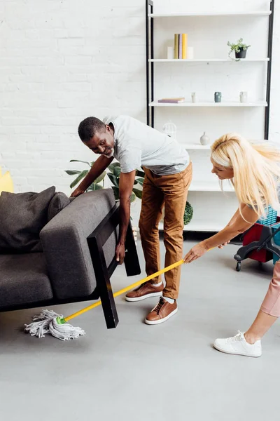Mulher loira limpeza chão com esfregona enquanto Africano americano levantar sofá pesado — Fotografia de Stock