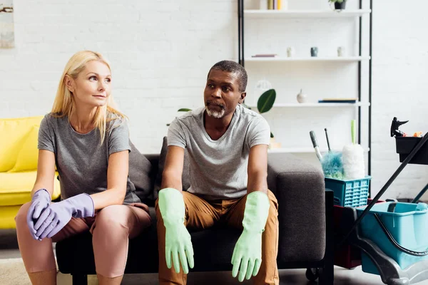 Tired couple in rubber gloves sitting on sofa after housecleaning — Stock Photo