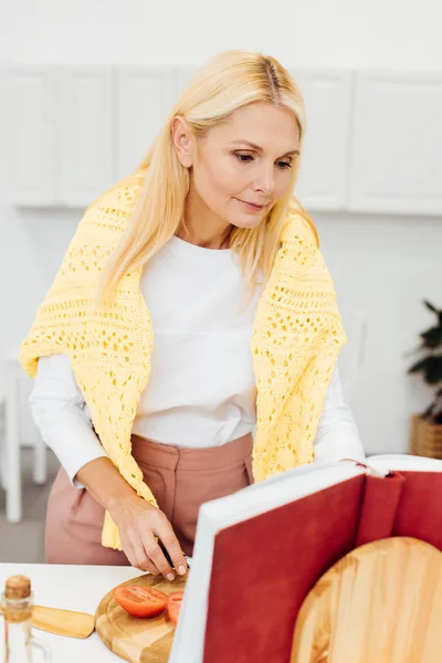 Beautiful mature woman cooking and reading recipe in cookbook at kitchen — Stock Photo