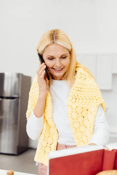 Donna sorridente che parla su smartphone e legge la ricetta nel libro di cucina in cucina — Foto stock