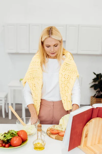 Beautiful blonde woman cooking salad with cookbook at kitchen — Stock Photo