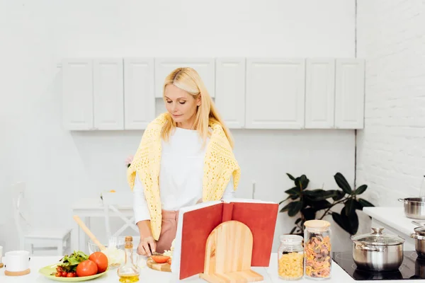 Beautiful blonde woman cooking salad with cookbook at kitchen — Stock Photo