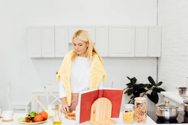 Bella donna bionda che cucina insalata con ricettario in cucina — Foto stock