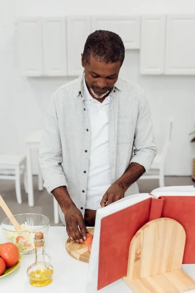 Uomo africano americano che cucina insalata con ricettario in cucina — Foto stock