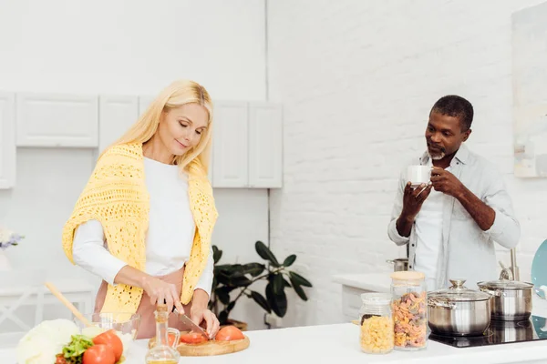 Reife blonde Frau kocht Abendessen während afrikanisch-amerikanischer Mann Kaffee trinkt — Stockfoto