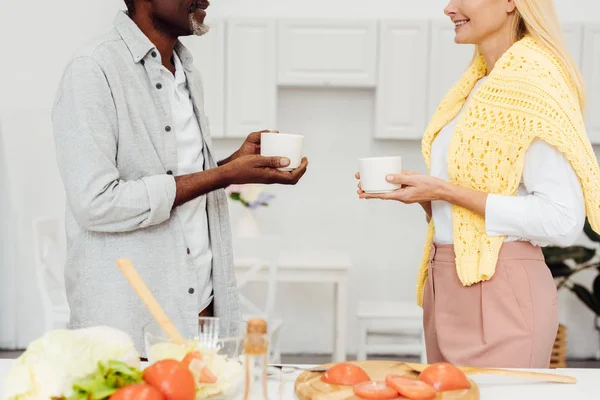 Cropped view of mature blonde woman and african american man drinking coffee together at kitchen — Stock Photo