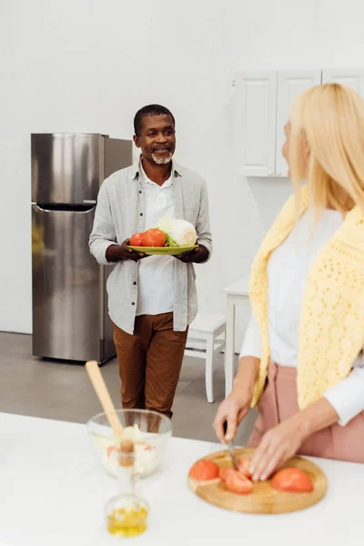 Frau schneidet Tomaten auf Schneidebrett, während afrikanisch-amerikanischer Mann Gemüse hält — Stockfoto