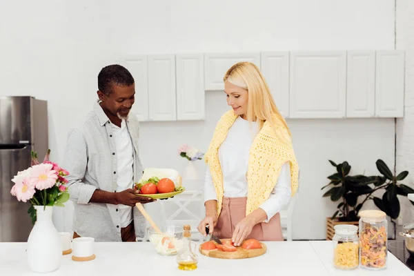 Зріла жінка нарізає помідори на дошці, афроамериканський чоловік тримає тарілку з овочами — стокове фото