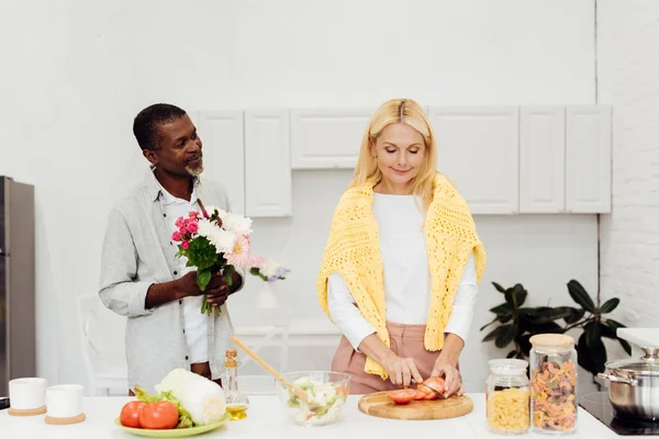 Bel homme afro-américain cadeaux fleurs à mature femme blonde à la cuisine — Photo de stock