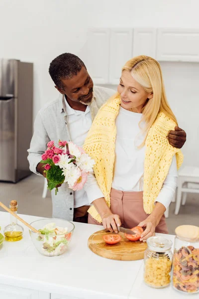 Guapo afroamericano marido regalando flores a madura rubia esposa en la cocina - foto de stock