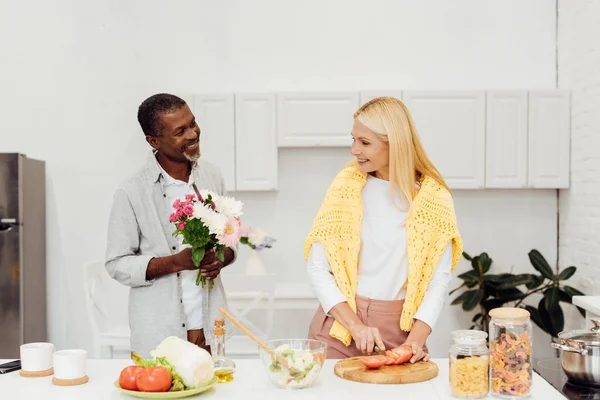 Guapo afroamericano marido regalando flores a madura rubia esposa en la cocina - foto de stock