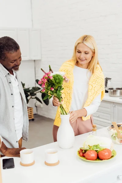 Bel homme afro-américain offrant des fleurs à une femme blonde mature — Photo de stock