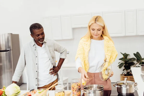 Africano americano hombre viendo rubia mujer cocinar cena - foto de stock