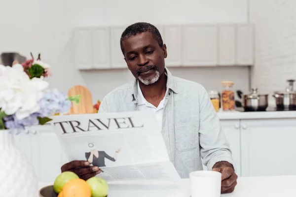 Hombre afroamericano sentado en la mesa y leyendo el periódico de viajes en la cocina - foto de stock