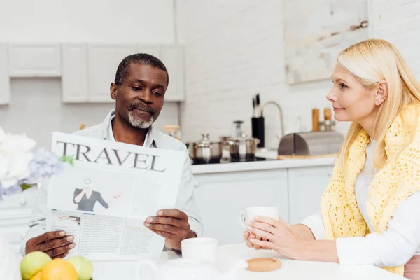 Afro-américain homme lecture journal tout en souriant femme mûre boire du thé — Photo de stock