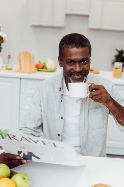 Homme afro-américain boire du café et lire un journal de voyage à la cuisine — Photo de stock