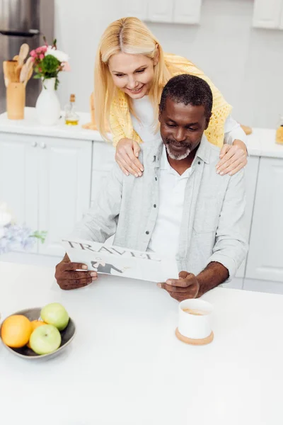 Mulher sorridente madura abraçando o homem enquanto lê jornal de viagem na cozinha — Fotografia de Stock