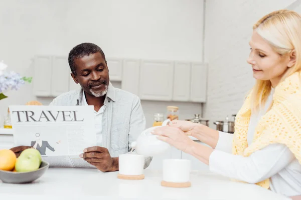 Africain américain lecture journal de voyage tandis que blonde femme mûre verser le thé dans des tasses — Photo de stock