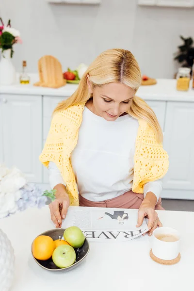 Attractive blonde mature woman reading travel newspaper at kitchen — Stock Photo