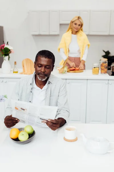 Homem americano africano ler jornal enquanto mulher cozinhar café da manhã — Fotografia de Stock