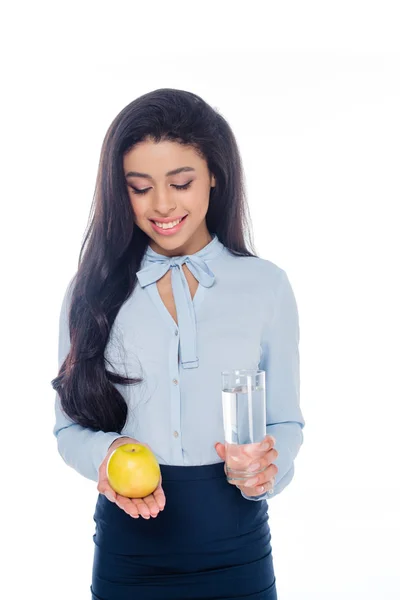 Souriante jeune femme afro-américaine tenant un verre d'eau et de pomme isolé sur blanc — Photo de stock