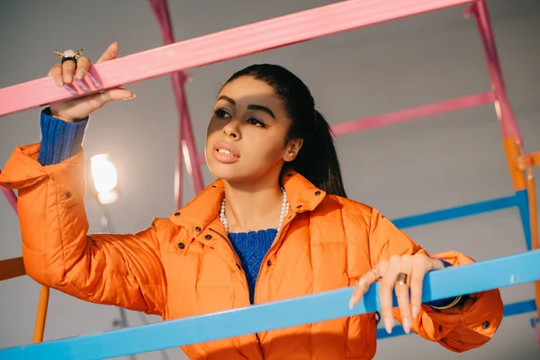 Belo modelo feminino afro-americano em casaco de inverno laranja posando perto de andaime colorido em estúdio com holofotes — Fotografia de Stock