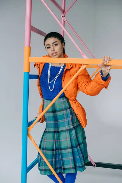 African american girl in orange winter jacket and checkered skirt posing near colorful scaffold in studio — Stock Photo