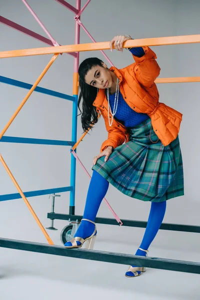 Stylish girl in orange winter jacket, checkered skirt and blue tights posing near colorful scaffold in studio — Stock Photo