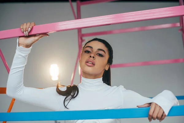 Portrait of african american female model in white turtle neck posing near scaffold in studio with spotlight — Stock Photo