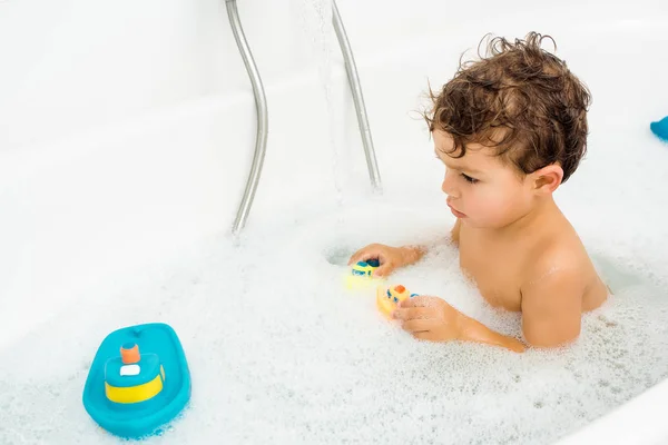 Маленький мальчик играет с игрушками для ванны в белой ванной — стоковое фото