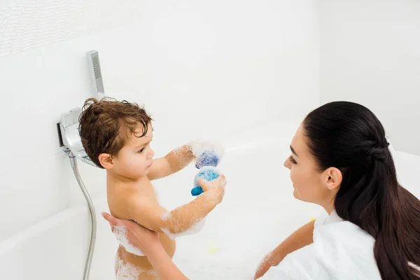 Filho mostrando brinquedos para morena mãe no banheiro branco — Fotografia de Stock