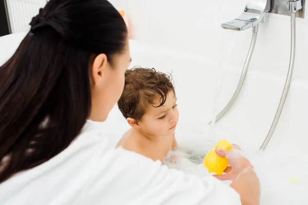 Morena mujer mostrando amarillo pato juguete a hijo en baño - foto de stock