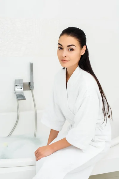Schöne Frau sitzt im weißen Bademantel im weißen Badezimmer — Stockfoto