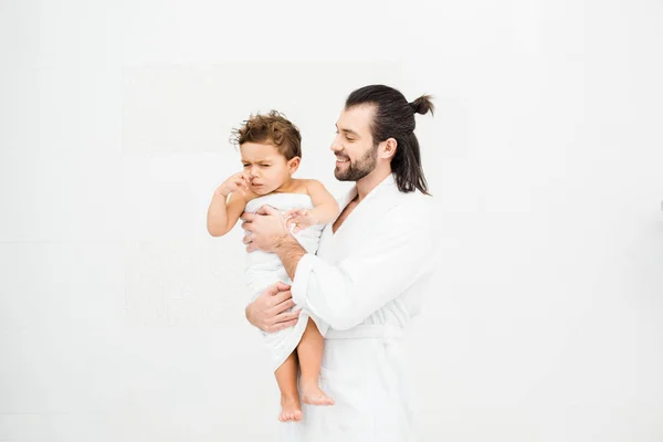 Father in bathrobe looking at son in towel and smiling on white — Stock Photo