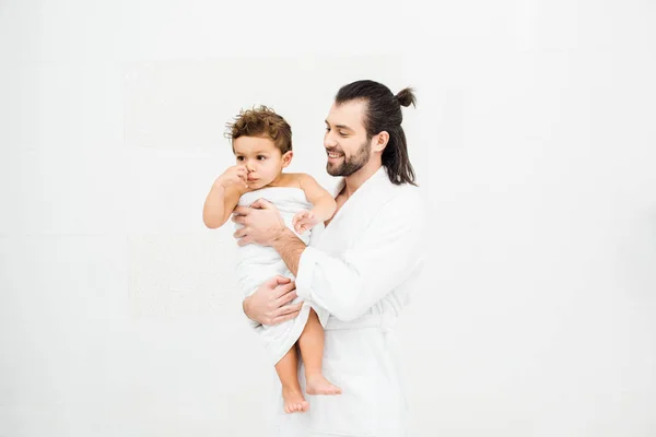 Papá en albornoz sonriendo a su hijo pequeño en toalla en blanco - foto de stock