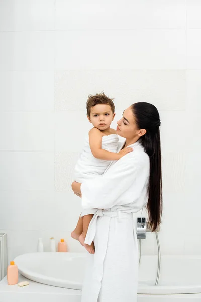 Mother holding and looking at toddler son in white towel in bathroom — Stock Photo